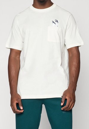 Mand iført hvid T-shirt med korte ærmer og lille lomme på venstre bryst, der holder et par hvide og sorte stribede sokker.