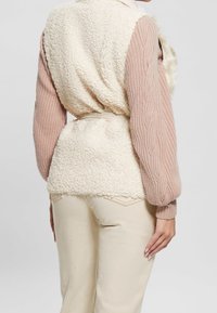 Vista posteriore di una persona che indossa un gilet in sherpa color crema sopra un maglione a trecce rosa pallido e pantaloni alti in crema, in piedi su uno sfondo bianco.