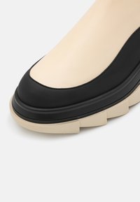 Gros plan d'une chaussure beige et noire à talon bas avec une semelle beige épaisse et segmentée sur fond blanc.
