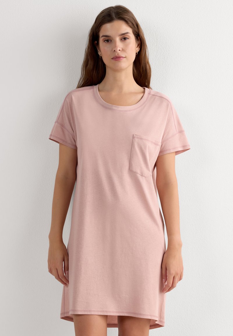 Mujer con cabello largo y castaño, que lleva un vestido corto de manga corta de color rosa claro con bolsillo en el pecho, de pie junto a una pared blanca.