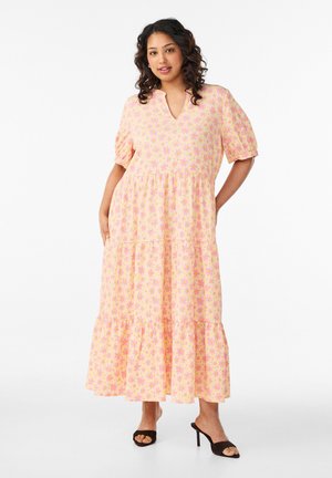 Naine kannab kollast keskmise pikkusega kleiti roosa lillemustriga, lühikeste pufvarrukatega, V-kaelusega ja mustade avatud varbaga kontsakingadega, seisab valge tausta ees.