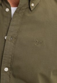 Camisa de botón verde oliva con cuello, que presenta un pequeño logo de ala bordado en el pecho y botones blancos. Textura suave.