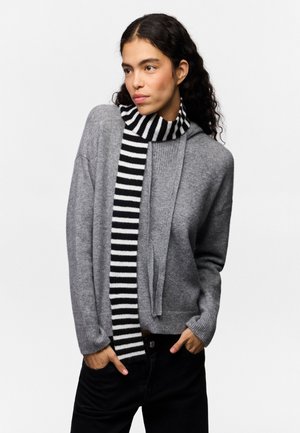 Kvinde med krøllet mørkt hår iført en grå sweater og sort-hvidstribet tørklæde, stående med hænderne i lommerne mod en hvid baggrund.