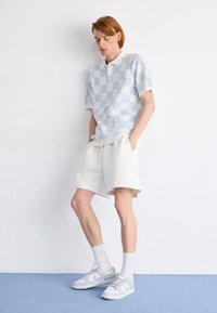 Ljusblå kortärmad polo med rutmönster, vit krage; matchad med krämfärgade shorts och grå sneakers med ljusgrå detaljer.