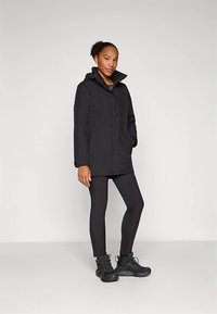 Chaqueta impermeable negra con capucha, cremallera frontal y bolsillos laterales, combinada con mallas negras y calzado negro resistente.