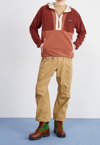 Felpa con cappuccio marrone in pile con un pannello crema, abbinata a pantaloni cargo khaki e stivali marroni con accenti verdi in peluche e lacci rossi.