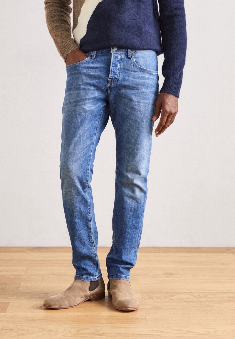 Světle modré džíny se slim fit střihem, s kapsami a jemným vyblednutím, spárovány s béžovými semišovými kotníkovými botami na dřevěné podlaze.