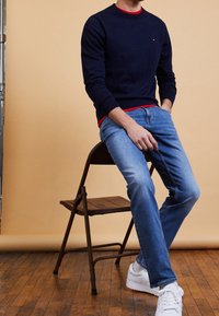 Suéter de cuello redondo azul marino sobre una camisa roja, combinado con jeans azul claro y zapatillas blancas. La persona está sentada en una silla plegable.