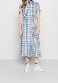 Žena v modro-bílých kostkovaných midi šatech s páskem na zavázání, bílých teniskách, kostkovaných ponožkách a šátku na krku, stojící před jednobarevným pozadím.