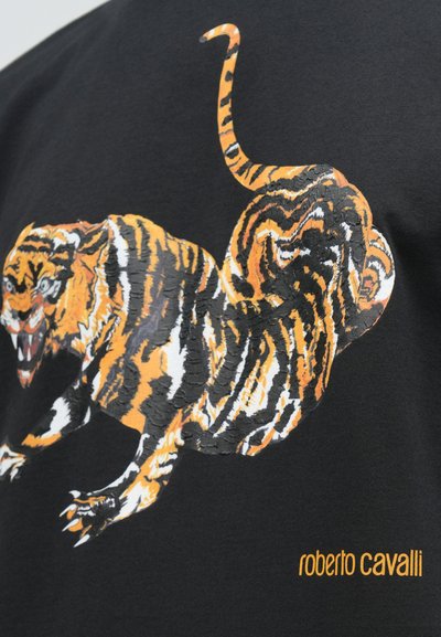 Tigra ilustrācija ar oranžiem, melniem un baltiem svītrainiem, kas iznāk uz melnas auduma, zem tā oranžā krāsā ir uzraksts "roberto cavalli".