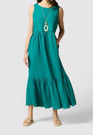 Woman wearing sleeveless teal maxi dress with gathered hem, beige flat sandals, and long pendant necklace, standing with one hand in pocket.