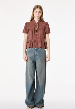 Jeune femme aux cheveux longs portant de grandes boucles d’oreilles, un chemisier marron boutonné avec des manches bouffantes et un jean bleu délavé à jambes larges, debout.