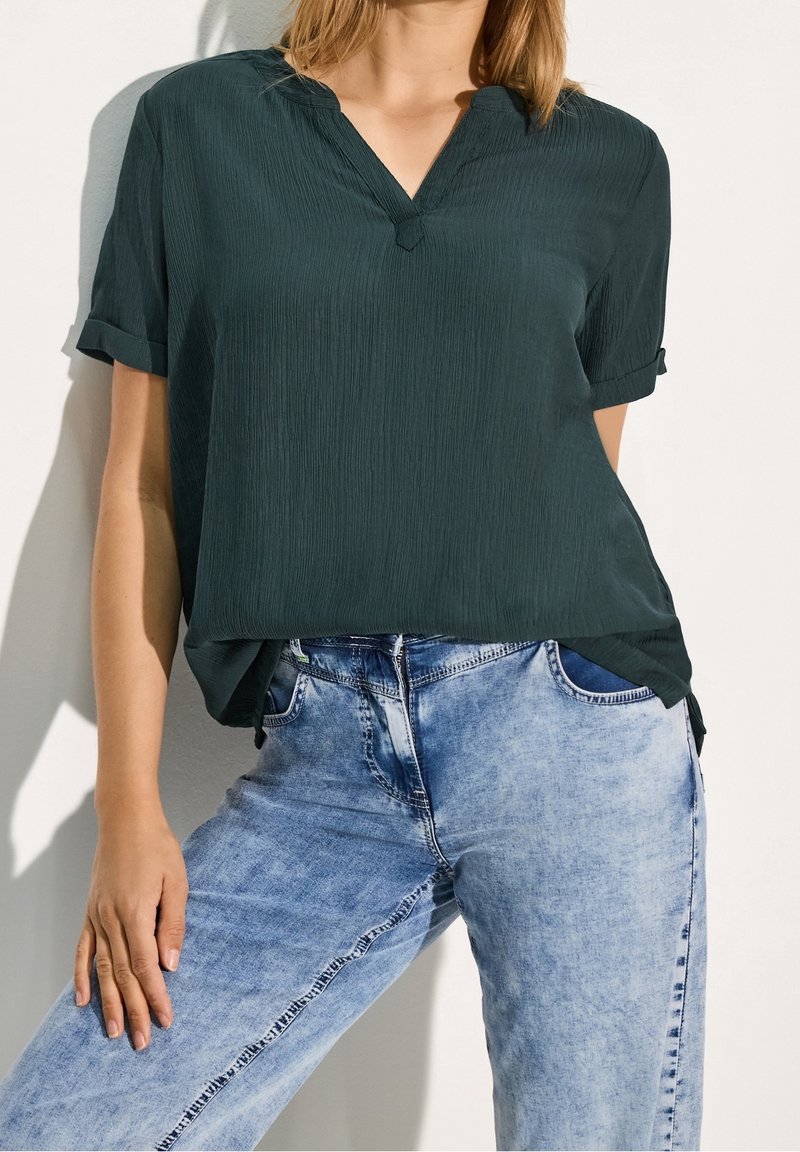 Femme portant un chemisier vert foncé à manches courtes, rentré dans un jean taille haute bleu clair, posant avec une main sur la cuisse.