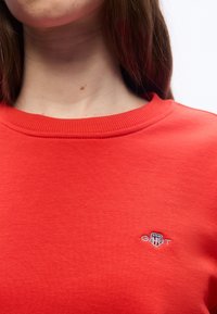 Κοντινό πλάνο ενός ατόμου που φοράει φωτεινό κόκκινο φούτερ GANT με έν α μικρό κεντημένο λογότυπο κοντά στην κλείδα.