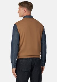 Rudas megztas liemuo užmestas ant mėlynos džinsinės ilgomis rankovėmis marškinių, turinčių paprastą dizainą ir raukšlėtą apatinę dalį, derinamas su tamsiais džinsais.