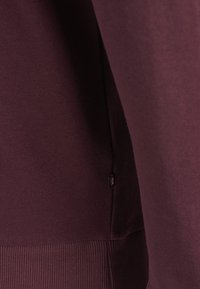 Bordeauxrode trui met gladde stof, geribbelde zoom en een klein logotagje in de zijnnaad, waarmee het eenvoudige en moderne ontwerp wordt benadrukt.