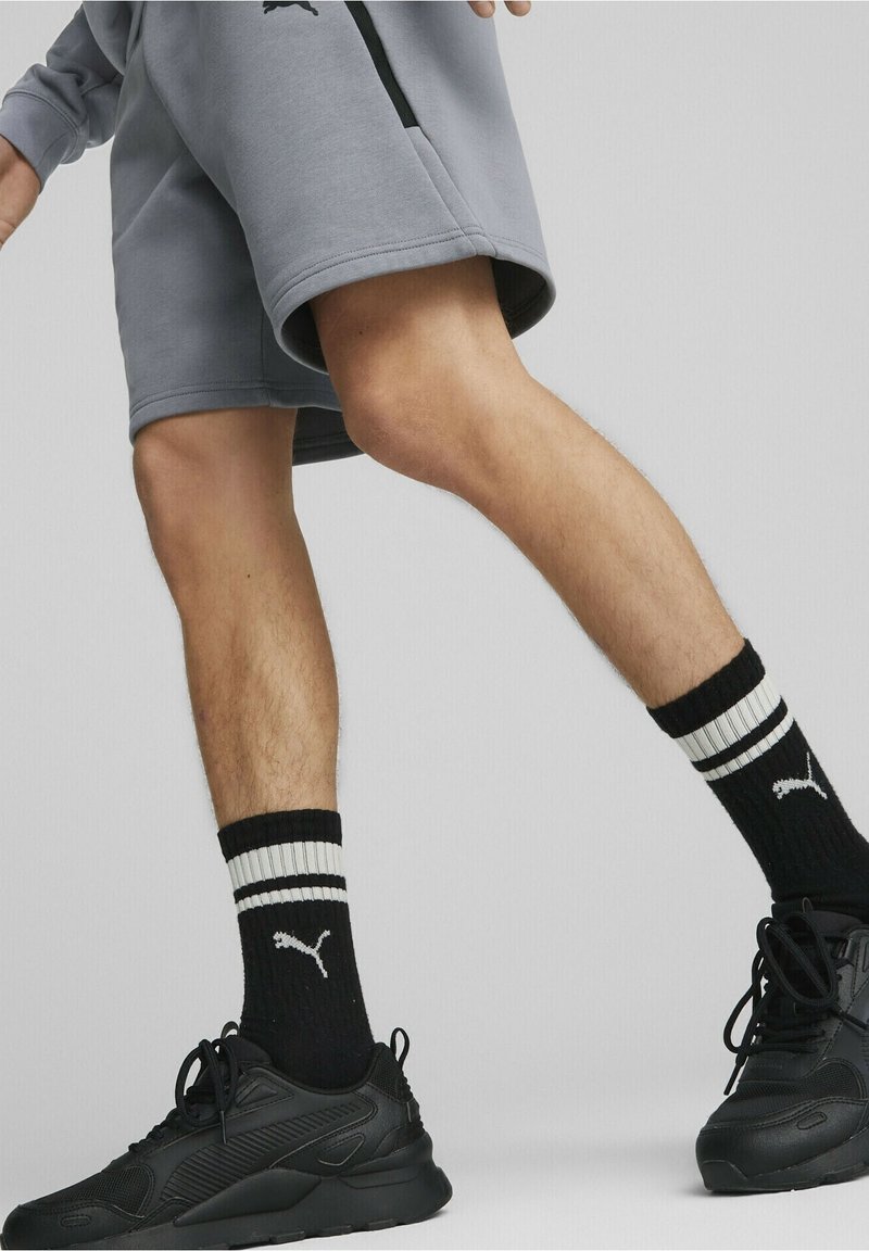 Personne portant un short gris, des chaussettes Puma noires et blanches, et des baskets Puma noires marchant sur un fond clair uniforme.
