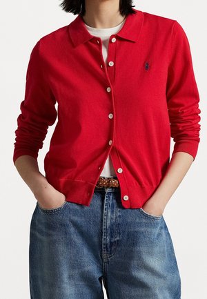 Personne portant un cardigan rouge boutonné par-dessus une chemise blanche avec un jean bleu et une ceinture tressée marron, les mains dans les poches.