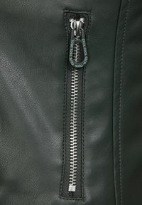Närbild av en svart läderjacka med en silvermetallblixtlås och en branding dragflik samt synliga sömdetaljer på en texturerad yta.