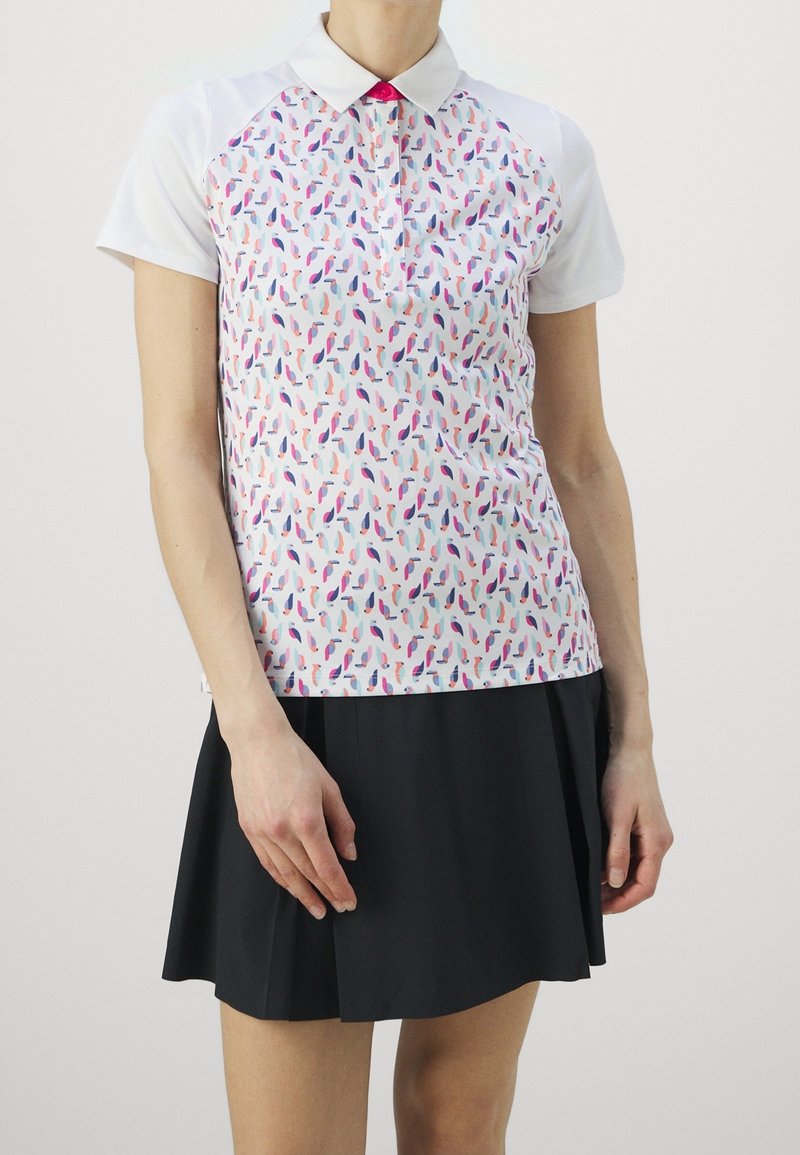 Femme portant un polo blanc à manches courtes avec des motifs d'oiseaux colorés et une jupe plissée noire, debout devant un fond blanc uni.