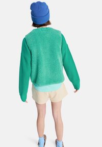 Zielona teksturowana kurtka polarowa z prążkowanymi rękawami, zestawiona z jasnoniebieskim topem, beżowymi szortami i niebieską dzianą czapką.