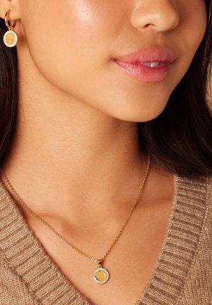 Collier en pendentif en or avec un charme circulaire présentant un design, posé sur un pull beige texturé. Les boucles d'oreilles présentent un design similaire.