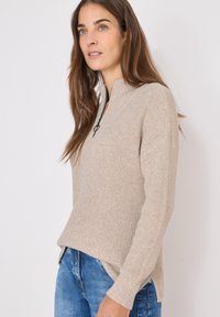 Beżowy sweter z prążkowanej dzianiny o wysokim dekolcie i pół zamkiem błyskawicznym. Posiada długie rękawy i boczne rozcięcia na dolnym brzegu. Wygodny splot dzianiny.