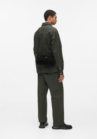 Borsa a tracolla nera con dettaglio in rete testurizzata, dotata di scomparto con zip e tracolla regolabile. Il modello indossa un outfit verde oliva.