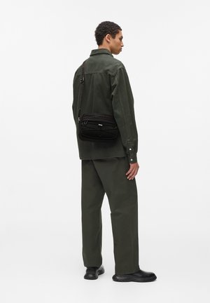 Schwarze Umhängetasche mit strukturiertem Mesh-Detail, ausgestattet mit einem Reißverschlussfach und verstellbarem Tragegurt. Das Modell trägt ein olivgrünes Outfit.