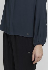 Blusa azul marino con mangas largas y abullonadas, que presenta una textura suave y un detalle de botón plateado en el dobladillo, combinada con pantalones oscuros.