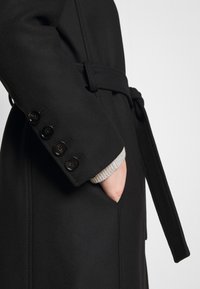 Sort frakke med en struktureret finish, der har et bælte, knappede manchet detaljer og en lomme. Den synlige ærme har en lys grå strikket foer.