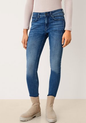 Person iført tætsiddende blå jeans og beige ankelstøvler står mod en ensfarvet baggrund.