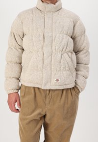 Beige puffjacka med hög krage, quiltat mönster och elastiska ärmslut. Har två sidofickor och ett litet logomärke vid fållen.