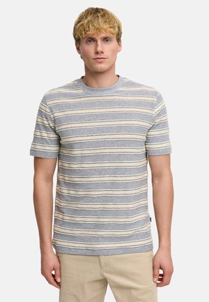 Junger Mann mit kurzem blondem Haar, der ein graues kurzärmeliges Hemd mit horizontalen weißen und gelben Streifen und beigen Hosen trägt.