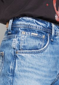Jeans in denim azzurro chiaro con trama consumata, cuciture visibili e una piccola etichetta con marchio sulla tasca posteriore con dettaglio a zip.