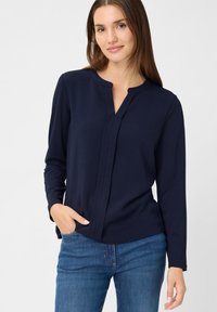 Blusa lunga a maniche di colore blu navy con colletto alla mandarino, patta frontale e una texture liscia, abbinata a jeans azzurri.