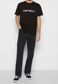 Mann in einem schwarzen Carhartt T-Shirt, dunkelgrauen geraden Jeans und schwarz-weißen Turnschuhen, steht mit einer Hand in der Tasche.