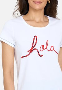 Camiseta blanca de manga corta con el texto bordado en rojo y azul marino "hola", cuello redondo y sutiles detalles de costura.