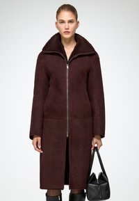 Burgundia värvi shearling-jope kõrge kraega, täispikk eeslukk, pikad varrukad ja sirge siluett, koos musta käekotiga.