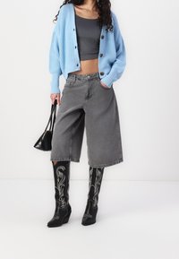 Kobieta ubrana w jasnoniebieski kardigan, ciemnoszare sukienkowe spodnie o szerokich nogawkach do połowy łydki, czarne wyszywane kozaki za kolano, trzymająca czarną torebkę.