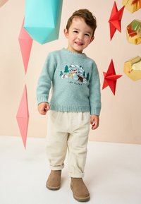 Pull en tricot turquoise avec broderie animale, pantalon crème à côtes, et bottines brunes. Décor géométrique en arrière-plan.