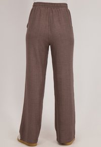 Losse, bruin gemêleerde broek met een elastische tailleband, gedragen door een staande persoon tegen een effen achtergrond.