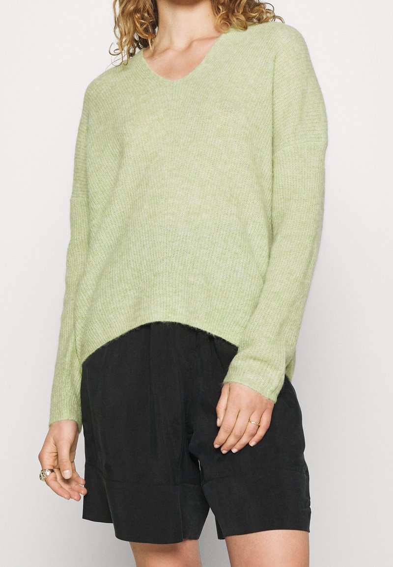 Femme portant un pull en tricot vert clair à col en V et un short noir ample, accessoirisé avec des bagues en or aux deux mains.