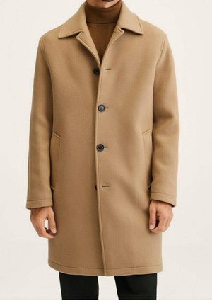 Homme portant un manteau camel longueur genoux avec des boutons noirs sur un pull à col roulé marron et un pantalon noir.