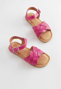 Sandali in pelle rosa con design a tracolla intrecciata, fibbia alla caviglia e sottopiede in suede marrone. Suola piatta con bordi arrotondati.