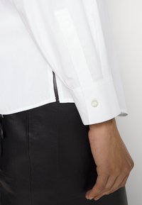 Camicia bianca a maniche lunghe con dettagli dei bottoni ai polsini, caratterizzata da un taglio dritto e fessure laterali, abbinata a pantaloni in pelle nera. Texture liscia.