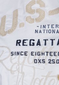 Biały materiał z haftowanym tekstem w kolorze beżowym i ciemnoniebieskim. Tekst zawiera "U.S. International Regatta Since Eighteen" oraz inne szczegóły.