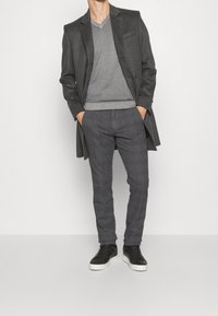 Šedý kabát přes šedý svetr s výstřihem do V, zkombinovaný se strukturovanými tmavě šedými kalhotami a černými teniskami s bílou podrážkou.