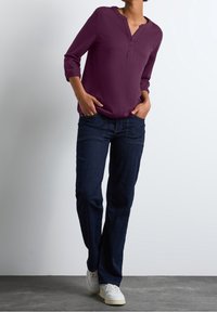 Woman wearing a long-sleeve purple blouse, dark blue jeans, and white sneakers standing with hands in pockets against a plain background.