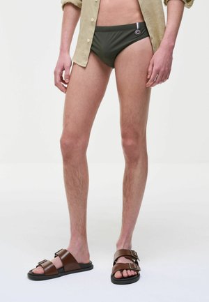 Uomo con camicia beige aperta, costume da bagno verde oliva e sandali marroni con fibbia, in piedi davanti a uno sfondo bianco semplice.